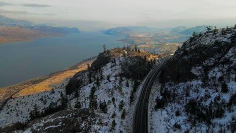 Reverse-Aerial-Shot-of-Highway-1-in-shade-running-through-rocky-hills-next-to-Kamloops-Lake-in-winter,-wonderful-golden-hour-light-over-the-partially-covered-desert-landscape