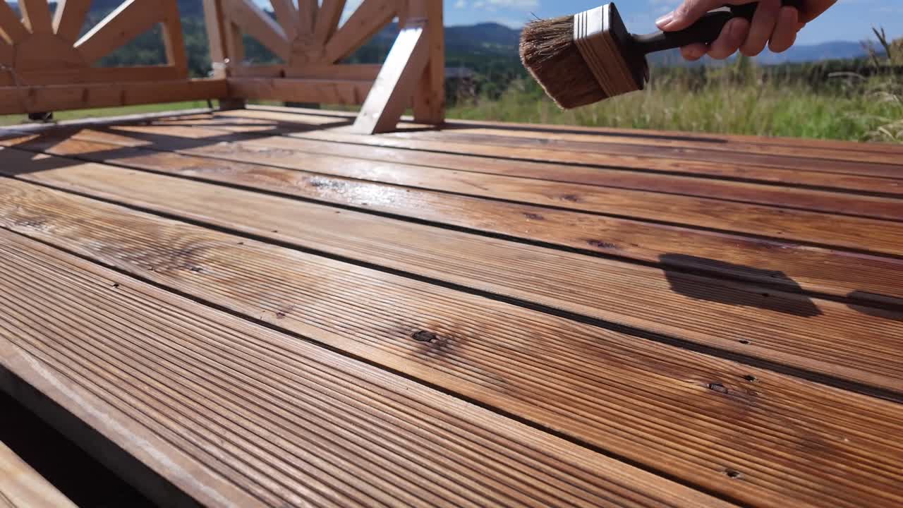 Working man brush strone and oils varnishing impregnating wooden terrace boards. Decorative railing, serene sky, clouds, and mountain horizon evoke home improvement and outdoor maintenance themes - 4K