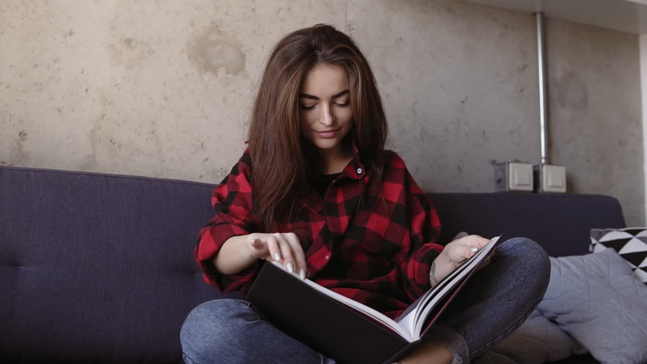 atractiva joven morena con cómoda camisa de franela roja sentada en un sofá y leyendo un libro. tiempo de relajación. imágenes de slomotion.