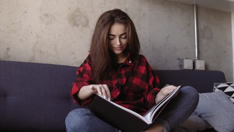 Attractive-young-brunette-girl-in-comfy-red-flannel-shirt-sitting-on-a-couch-and-reading-a-book.-Chilling-out-time.-Slomotion-footage.