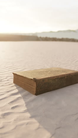 a weathered book lies open on a sandy beach with the sun setting in the distance