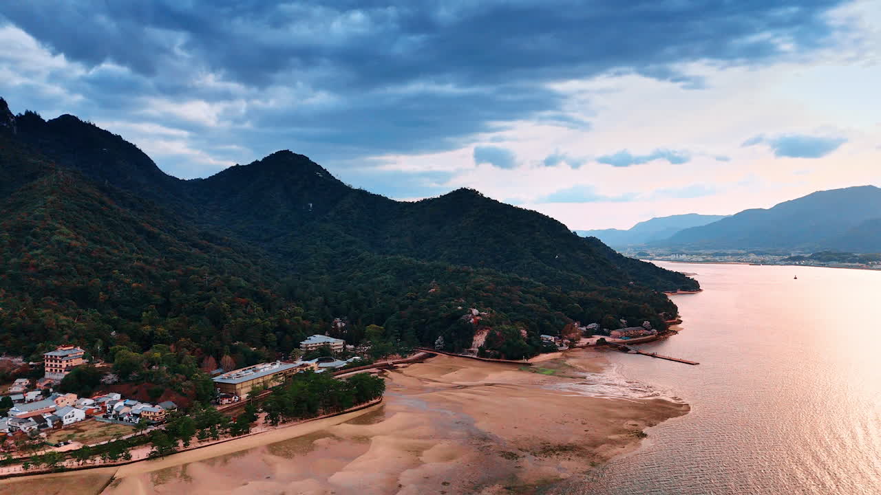 Sandy beach at the Inland Sea of Japan on the Miyajima island. Multiple houses are at the foot of the green mountain under the cloudy sky. Drone footage.