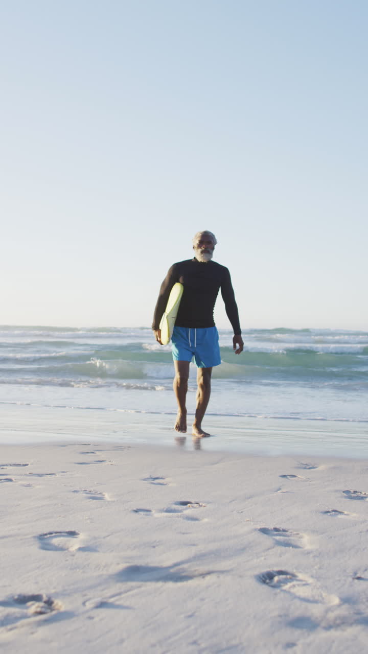 video vertical de un surfista afroamericano de edad avanzada caminando por la playa