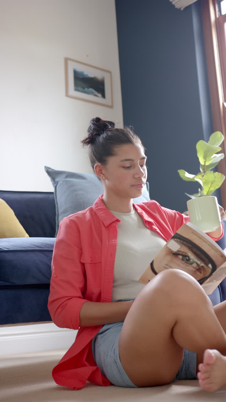 Vertical video of happy biracial teenage girl sitting on floor reading book with tea, slow motion