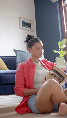 vertical video of happy biracial teenage girl sitting on floor reading book with tea, slow motion