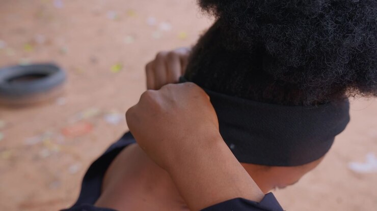 Close up shows woman with dark curly hair wearing black headband seen from side as they adjust their position. background is blurred but suggests outdoor environment with earthy tones