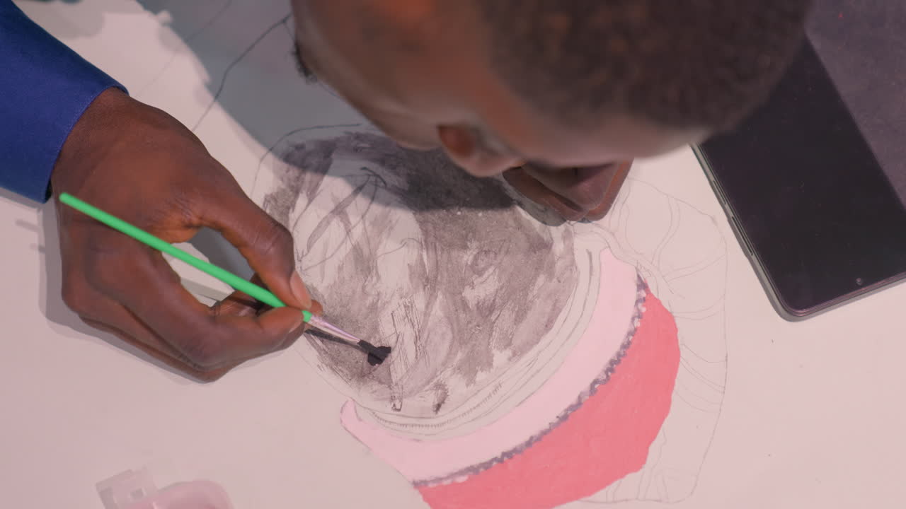 Overhead black artist painting black portrait with green brush, focused illustrator leaning over paper, careful tonal washes and charcoal lines build expressive face, studio table with phone