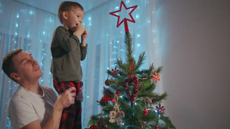 Papa-Hob-Den-Jungen-Auf-Die-Spitze-Des-Weihnachtsbaums,-Um-Einen-Weihnachtsstern-Darauf-Zu-Platzieren.-Vater-Und-Sohn-Schmücken-Gemeinsam-Den-Weihnachtsbaum.-Hochwertiges-4K-Filmmaterial