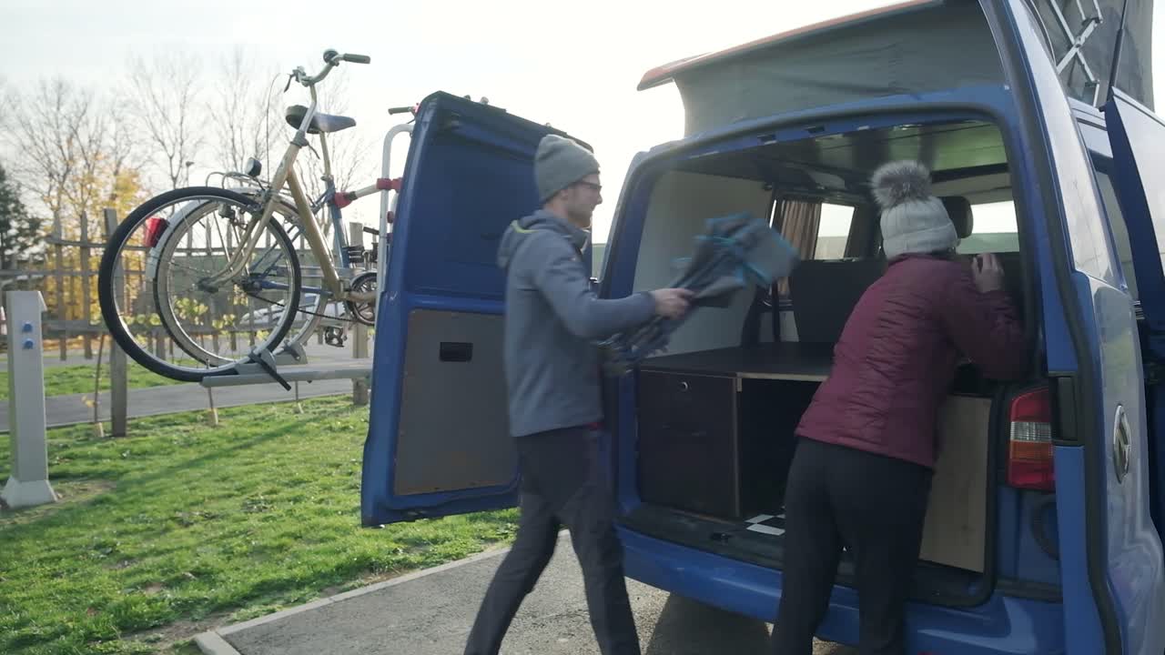A young couple packing up a chair and frisbee in the back of a blue campervan. On holiday on a sunny day playing in the field. A couple travelling in a campervan