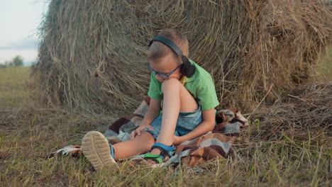 boy wearing headphones and glasses, sitting on blanket, listening to music, focused, with head resting on knee and vast farmland in background