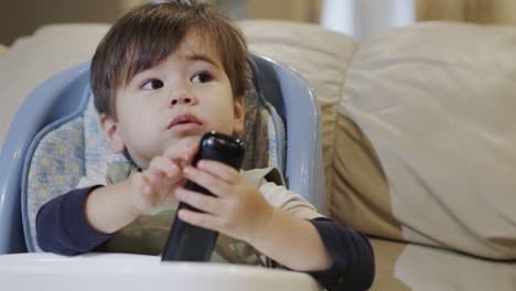 Cute-Asian-baby-sits-in-a-feeding-chair,-holds-a-remote-control-in-his-hands