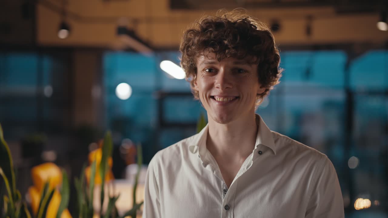 retrato de un joven feliz con el cabello rizado en una camisa blanca que está posando y sonriendo en la oficina de la noche