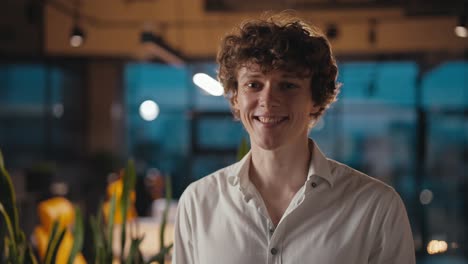 Portrait-of-a-happy-young-guy-with-curly-hair-in-a-white-shirt-who-is-posing-and-smiling-in-the-evening-office