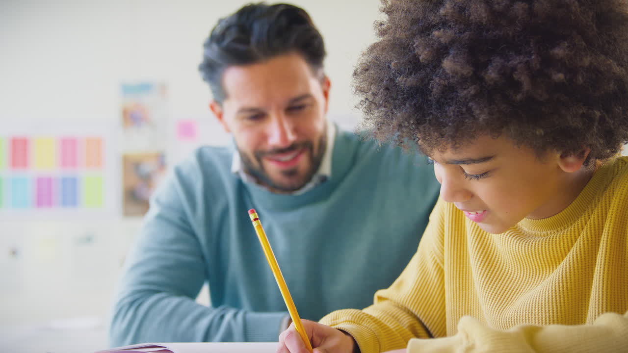 maestro masculino con estudiante masculino en el aula de la escuela sentado en el escritorio escribiendo en el libro juntos