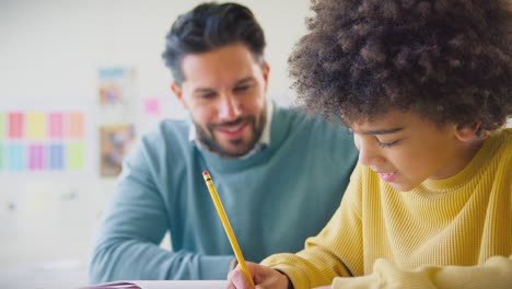 male teacher with male student in school classroom sitting at desk writing in book together