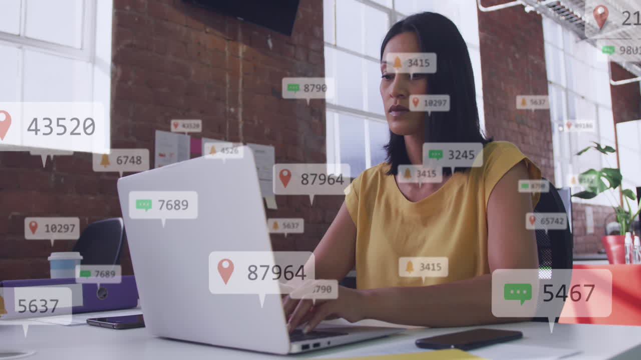 animación de iconos de las redes sociales flotando contra una mujer biracial usando una computadora portátil en la oficina