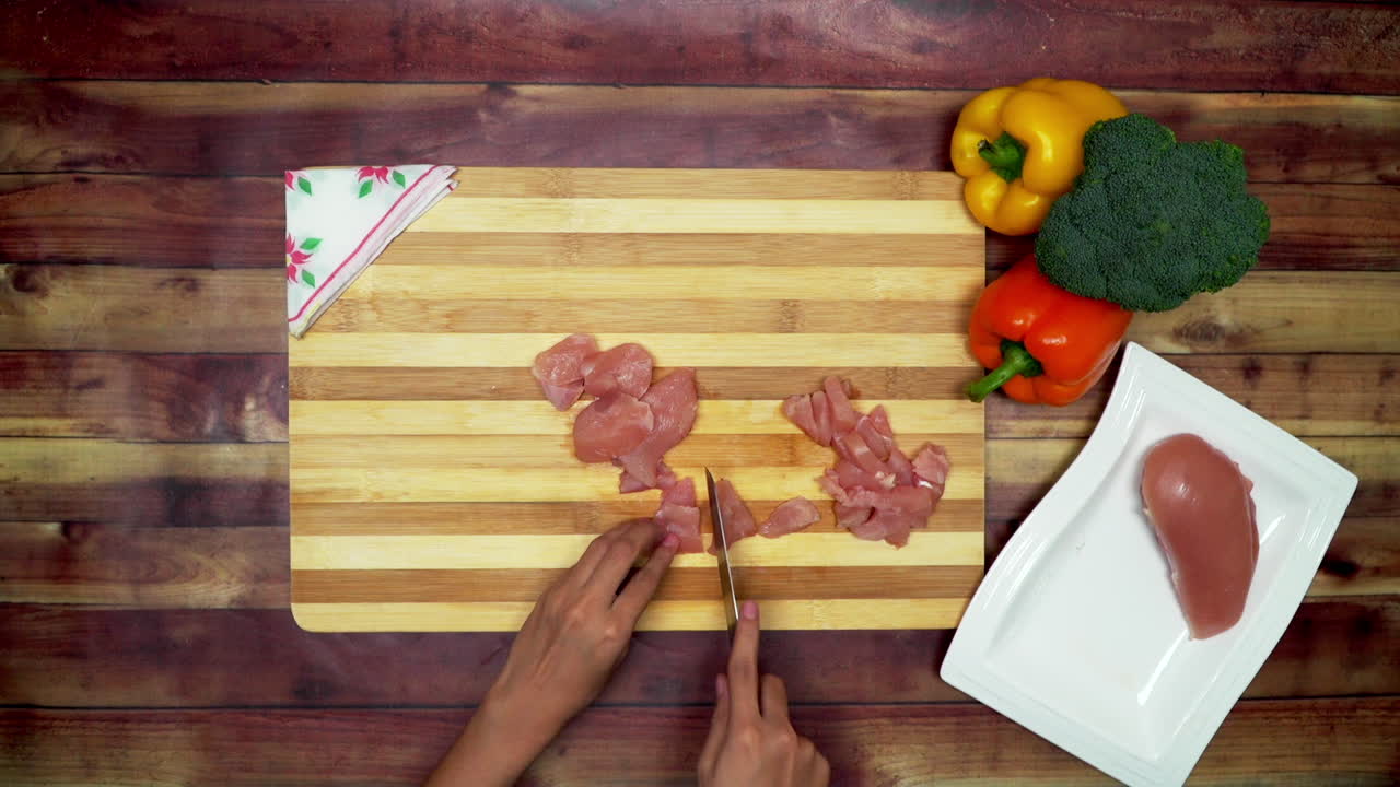 커팅 보드에 닭고기를 자르는 상위 뷰, 두 개의 큰 노란색 및 빨간색 고추, 뼈 없는 닭고기 조각, 강판 및 테이블에 있는 녹색 브로콜리