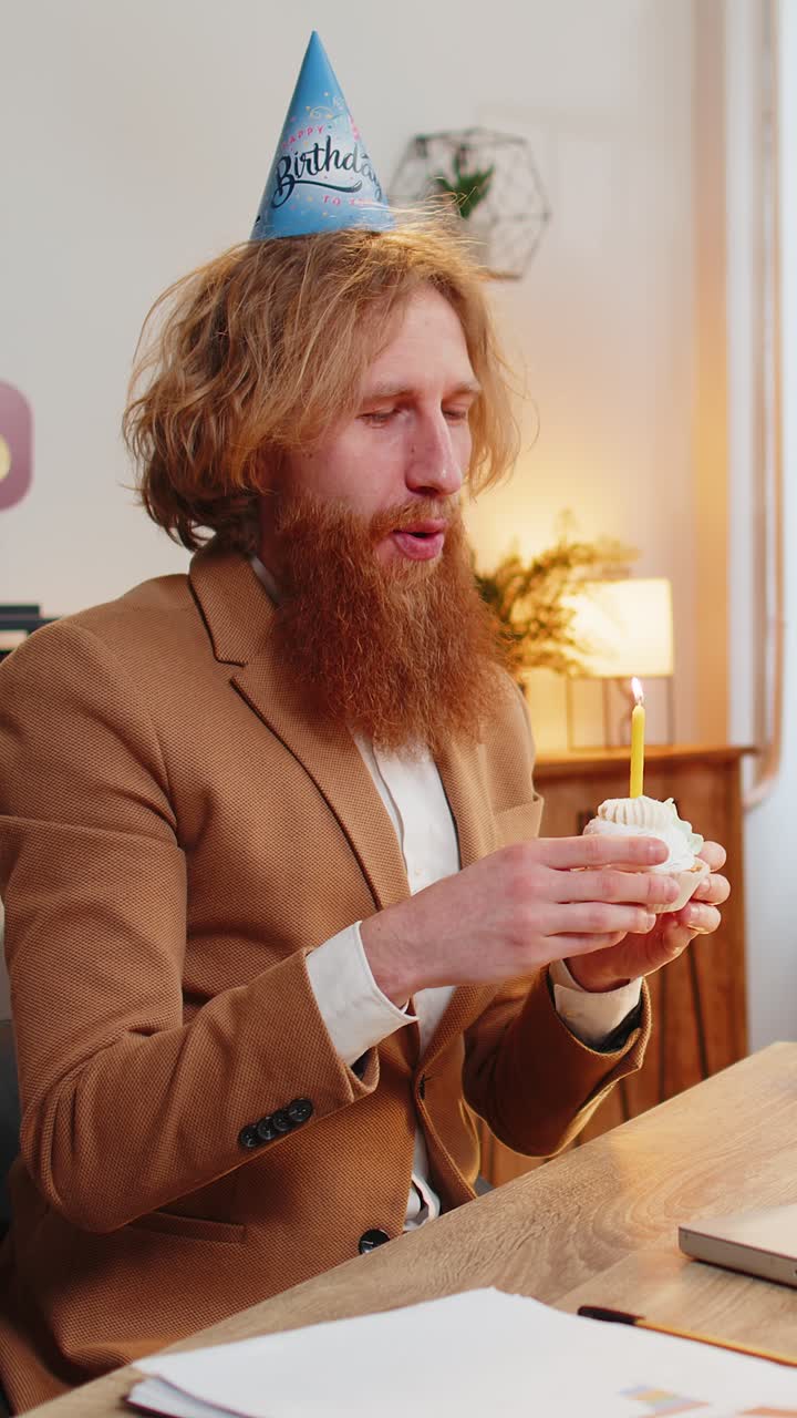 Un uomo d'ufficio felice indossa un cappello di compleanno festivo tiene un cupcake fa un desiderio soffia una candela accesa