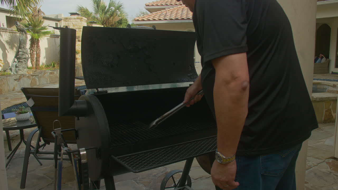 el hombre recoge las pinzas y mueve las rejillas en el patio trasero fumador de barbecue
