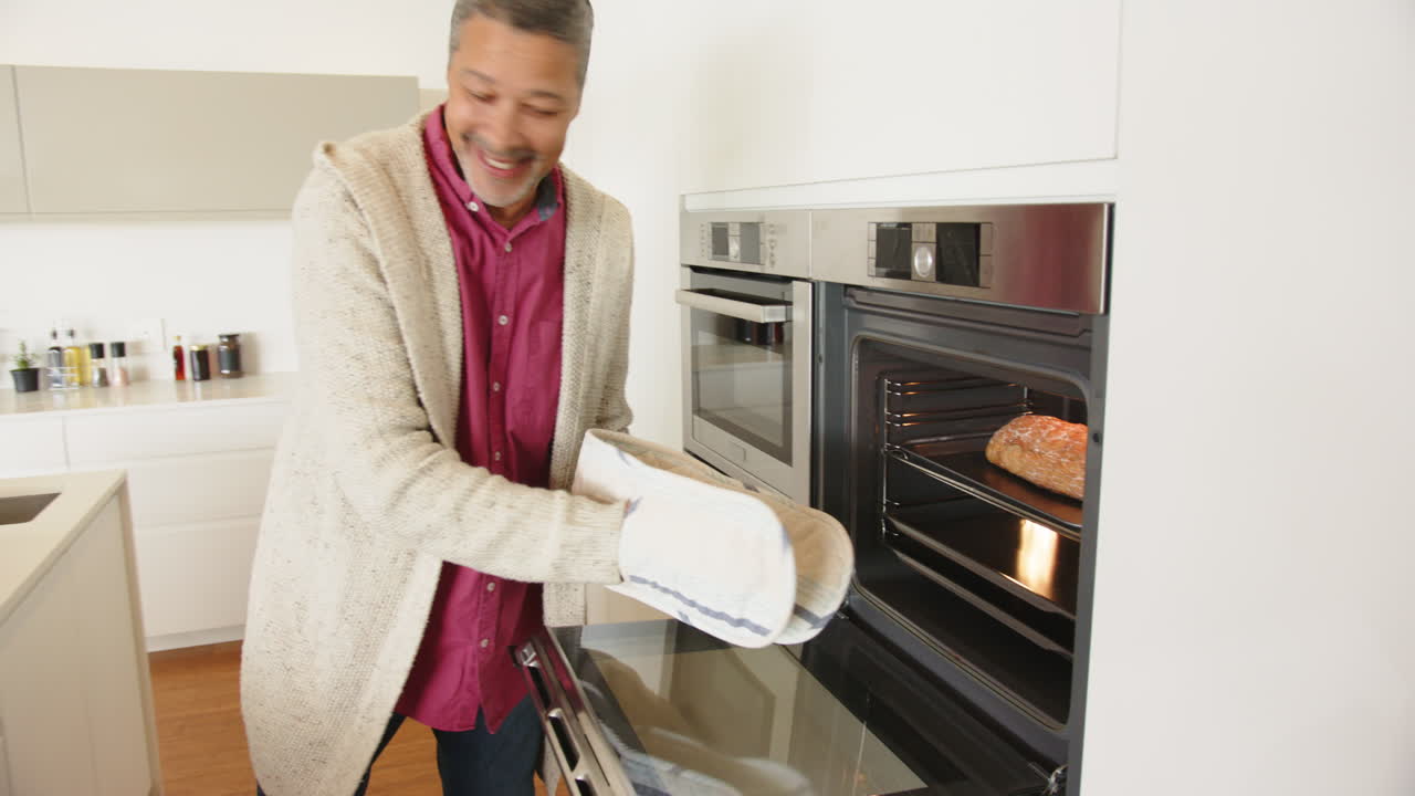 Uomo che usa guanti da forno, mettendo un piatto nel forno di una cucina moderna, sorridendo, a casa.
