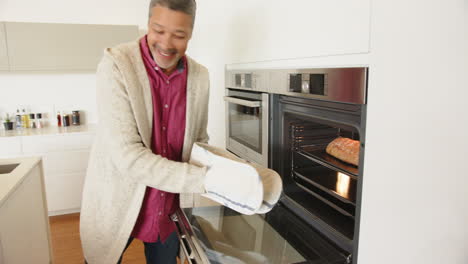using oven mitts, man placing dish in modern kitchen oven, smiling, at home