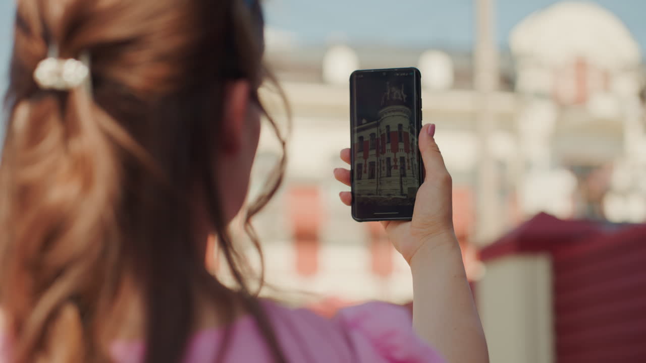 Jovem fotografando um edifício histórico ornamentado com um smartphone, rua da cidade ensolarada, close da tela do celular refletindo a fachada de tijolos vermelhos, cabelo solto preso com um clipe, blusa rosa, pouca profundidade de campo