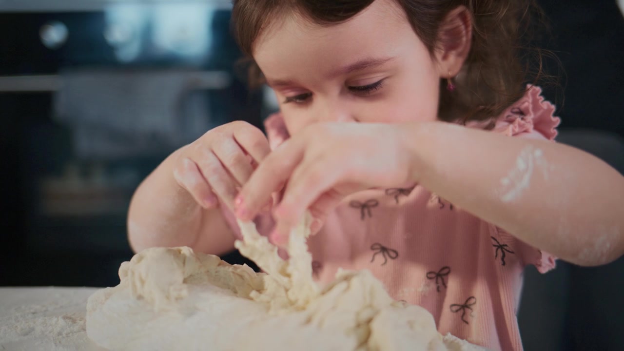 Young girl kneads dough with focused expression. She wears pink dress with bow pattern. Hands covered in flour show her playful effort. Scene captures joyful, domestic kitchen moment