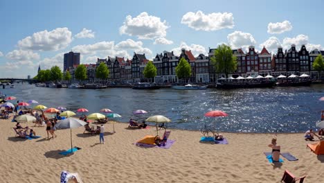 tourists relaxing on the beach in amsterdam, soaking up the sun under vibrant, colorful parasols while enjoying a picturesque view of the cityscape and the amstel river