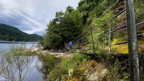 a hiker with a backpack walks across a suspension bridge in the wilderness next to a lake