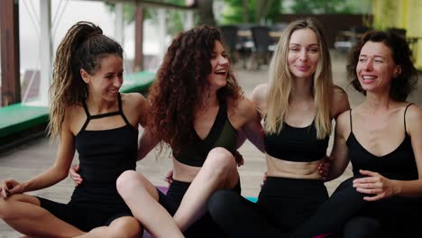 smiling girls in sportwear sit in an embrace on the floor, after practicing yoga