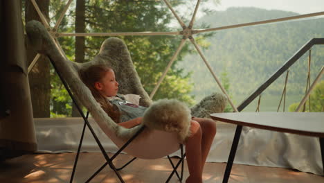 niño tranquilo sentado en un sillón de piel se relaja durante el día caluroso mientras observa paisajes de bosque de montaña. niña preescolar en glamping se esconde del calor al aire libre