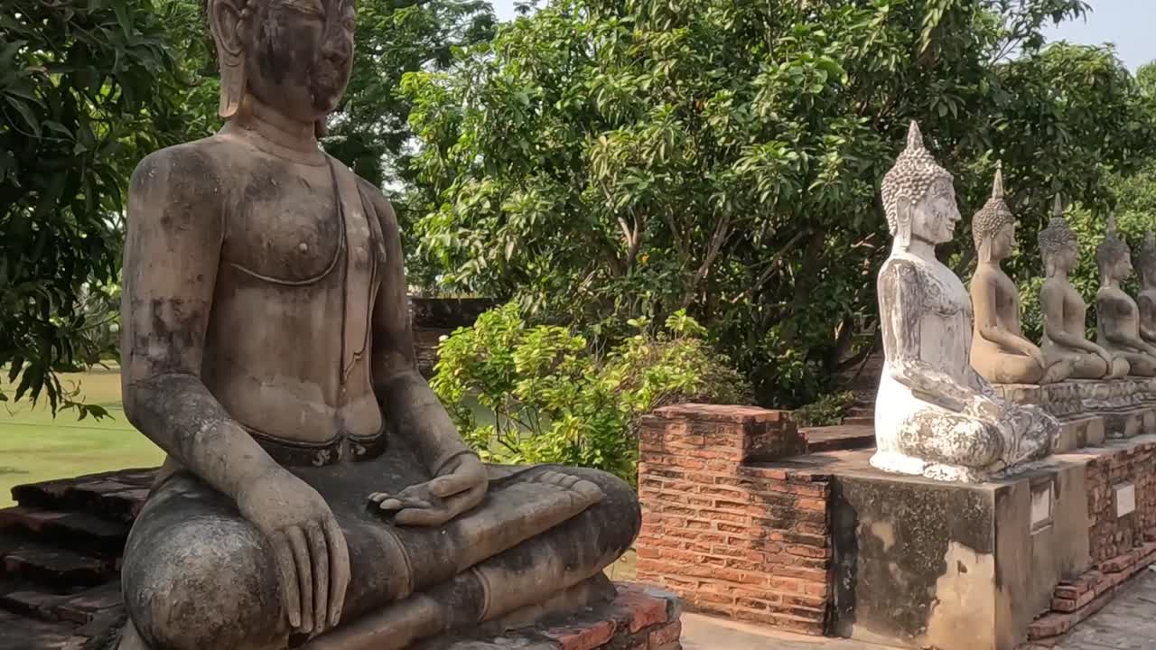 A series of weathered Buddha statues sit serenely on brick platforms surrounded by vibrant trees.