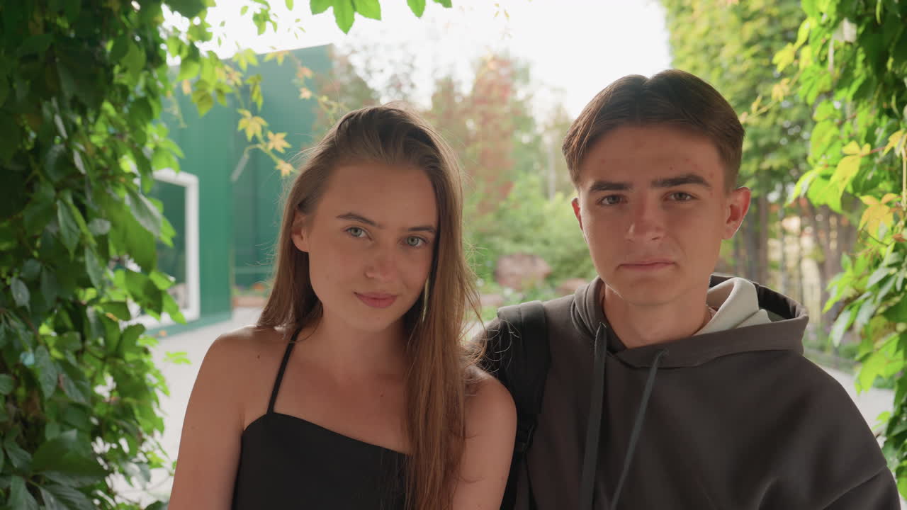 Adolescenti caucasici seduti sotto un arco di vite che si scambiano sguardi timidi, panchina del parco, cornice frondosa, luce solare soffusa, felpa casual e vestito estivo, sorrisi gentili e contatto visivo discreto che trasmettono una nuova amicizia