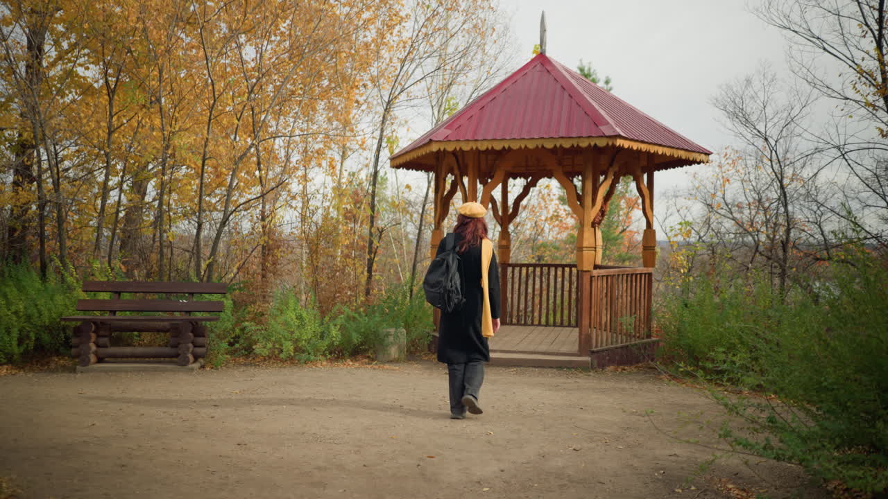 задний вид женщины в черном пальто, желтом шарфе и берете, идущей в деревянную беседку в спокойном парке, деревянная скамейка поблизости добавляет спокойной осенней обстановке, окруженной золотыми деревьями