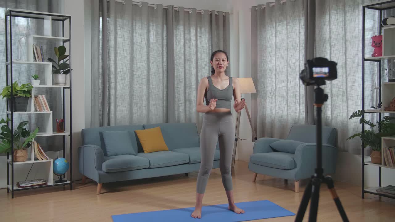 vista completa de joven entrenadora asiática mujer en ropa deportiva agitando la mano y hablando a la cámara mientras graba ejercicio de enseñanza en casa