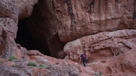 hiker approaching a large cave entrance in a red rock canyon