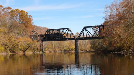 Train-crossing-bridge-over-river-during-autumn-from-the-viewpoint-of-a-boat-on-the-water-in-4K