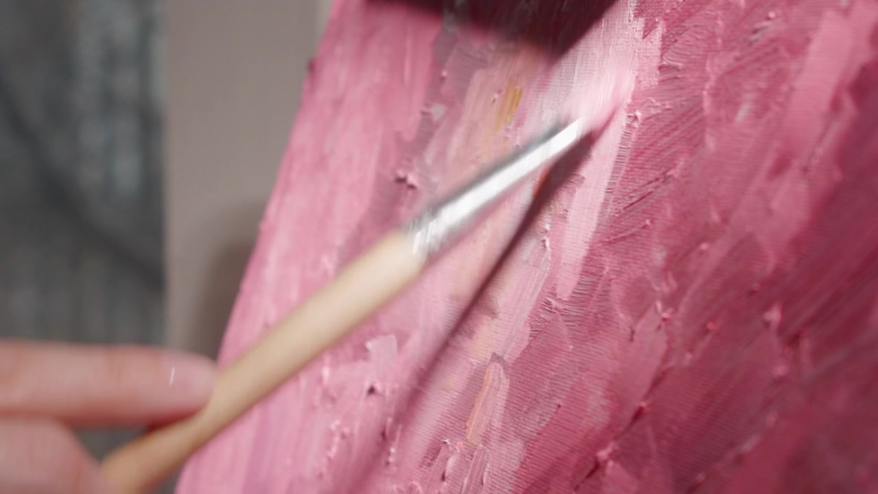 Primo piano di una mano che usa un pennello per applicare vernice rosa su una tela texturizzata, con una luce interna soffusa e uno sfondo sfocato che creano un'atmosfera creativa e calma.