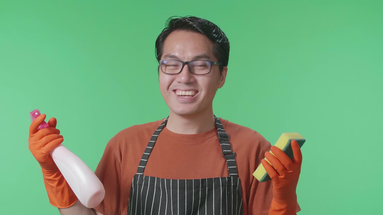 녹색 스크린 스튜디오에 서 있는 스프레이와 부 스폰지를 바라보는 아프론을 입은 미소 짓는 아시아 남성 집주인