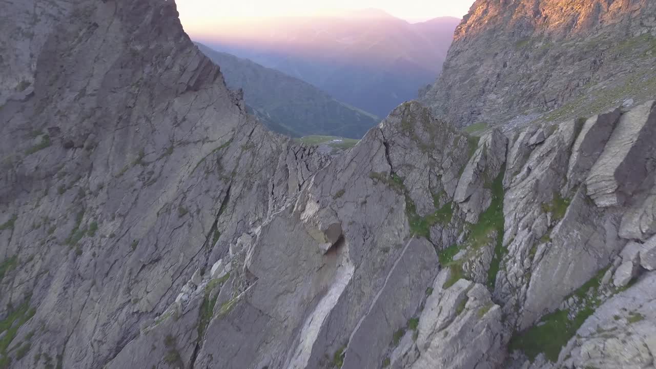 Vista drone che vola verso una cresta montuosa frastagliata in alto tra le montagne della Romania, con la luce colorata del tramonto sulle cime distanti e un lago che si rivela sotto