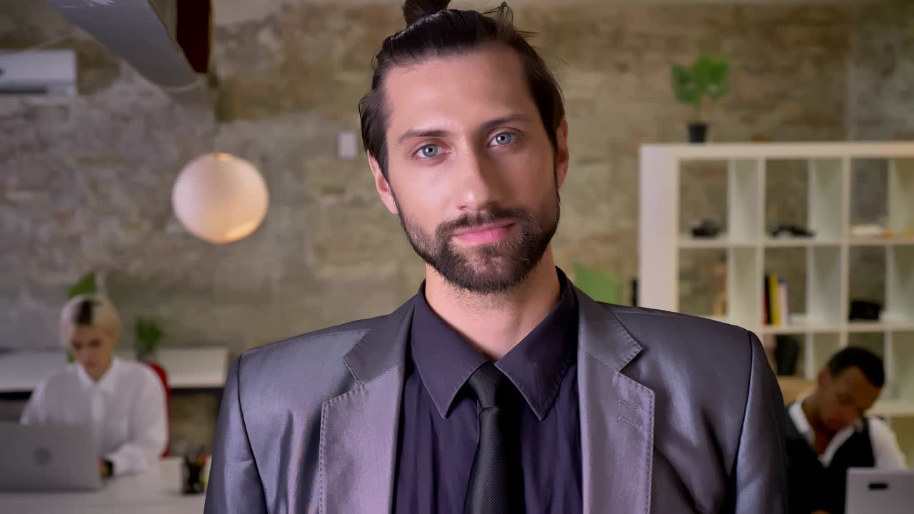 Handsome confident businessman with beard is watching at camera in office, colleagues are working with laptops, work concept