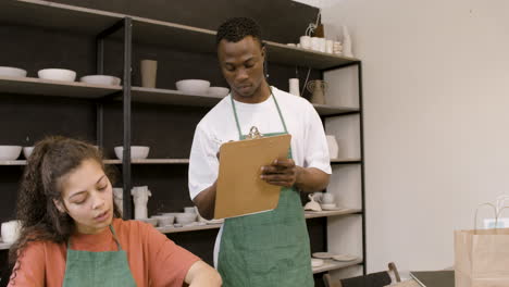 young woman wrapping handicraft ceramics with paper while her male colleague writing on clipboard in the pottery shop