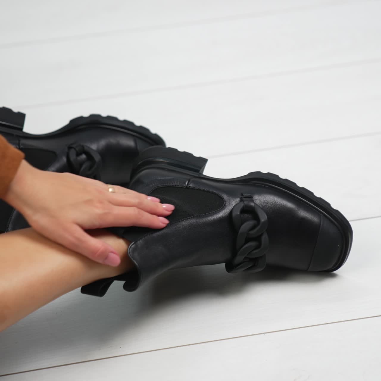 Square-toe black boots with chain decoration on the bare legs of a model. Girl demonstrates footwear sitting on the white floor