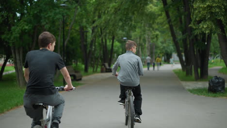 back view of two friends riding their bicycles on a park path with the one in grey top scratching his head, with a blurred view of people walking ahead, surrounded by lush green trees