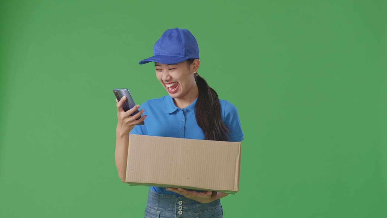 푸른 유니폼을 입은 아시아 여성 택배업자가 스마트폰을 보고 스튜디오에서 녹색 화면 배경에 상자를 배달하는 동안 성공을 축하하고 있습니다.