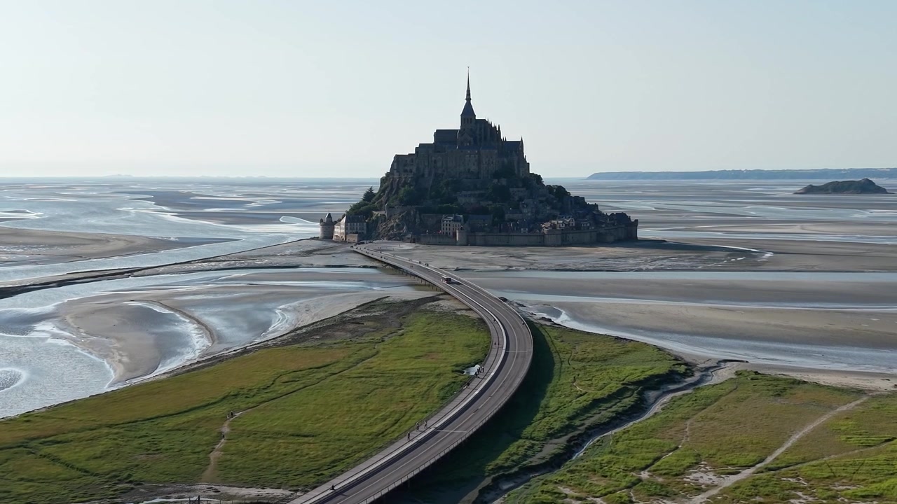 Vista aérea cinematográfica de drone do Mont Saint-Michel, apresentando a icônica abadia medieval conectada ao continente da Normandia pelo moderno aterro