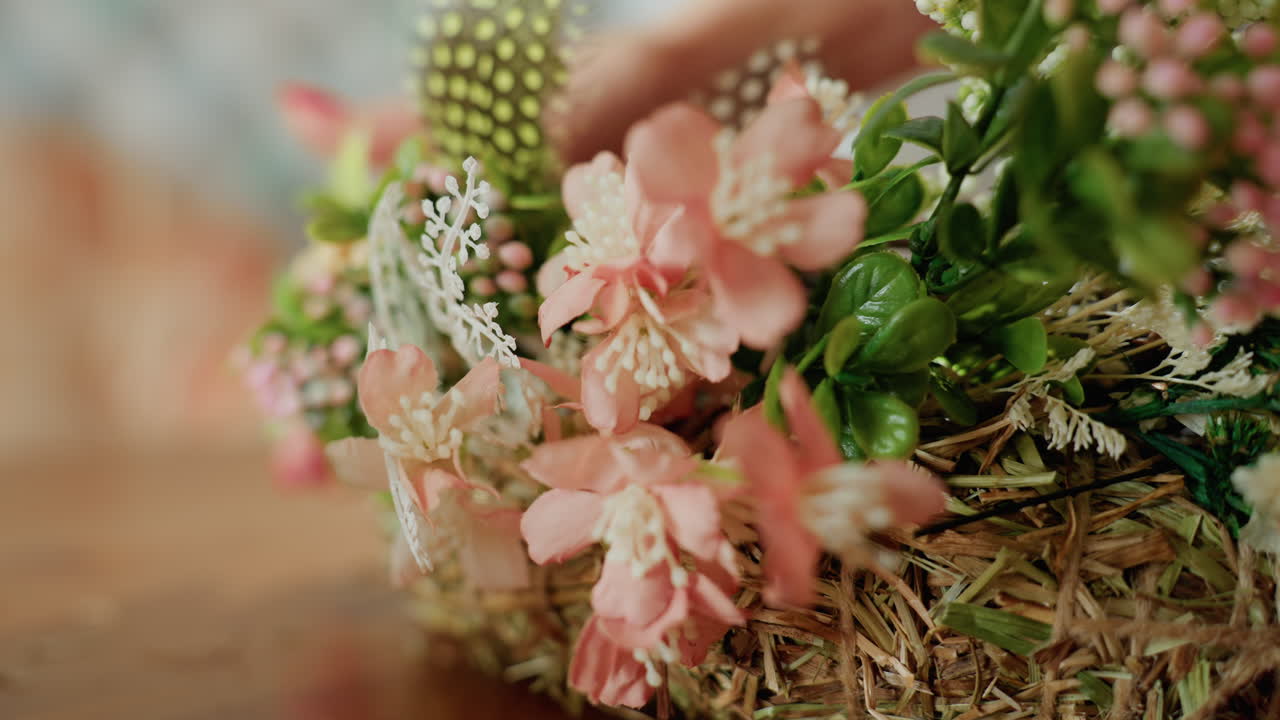 Close up female hand with red nails arranging handmade floral wreath decorated with pink blossoms, green leaves, berries, and dried flowers on straw base, highlighting delicate rustic craftsmanship