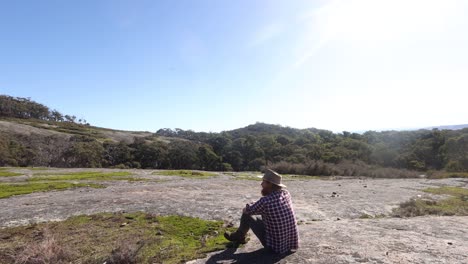 a bushman sits on a granite mountain and looks out accross the australian bush