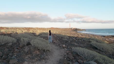 Woman-walking-through-path-next-to-coast-and-sea,-plants,-cactuses-with-lighthouse-in-background-stops-and-takes-a-picture,-Palm-Ar,-Tenerife,-Canary-Islands,-Spain