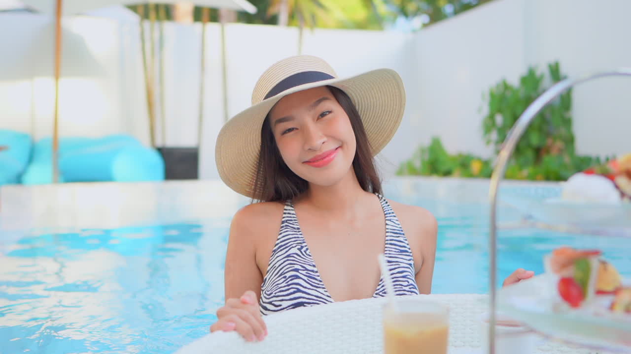Pretty young Asian woman pushing floating exotic breakfast tray full of fruits towards the camera in a swimming pool in the hotel resort
